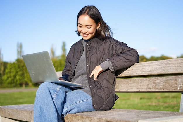 Porträt einer lächelnden Frau, die mit Laptop sitzt und an einem Projekt arbeitet oder aus der Ferne studiert und genießt, im Park zu sein