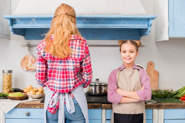 Porträt einer lächelnden Frau mit ihren Armen kreuzte das Betrachten der Kamera, die nahe der Mutter steht, die Lebensmittel in der Küche kocht