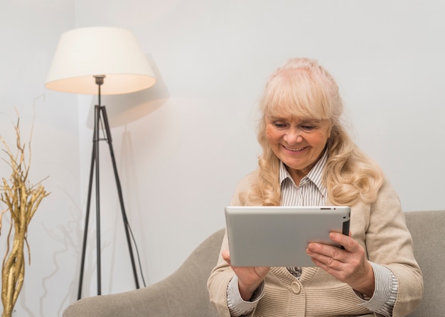 Porträt einer lächelnden Frau, welche die digitale Tablette sitzt auf Sofa betrachtet