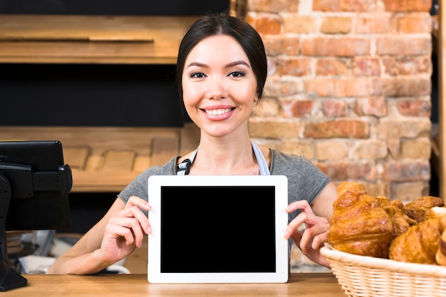 Porträt einer lächelnden jungen Frau, die digitale Tablette nahe dem Hörnchen auf Bäckereizähler zeigt