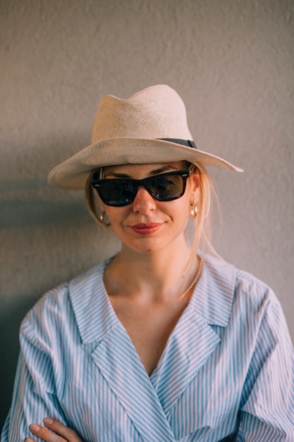 Kostenloses Foto porträt einer lächelnden jungen frau, die schwarze sonnenbrille und hut gegen wand trägt