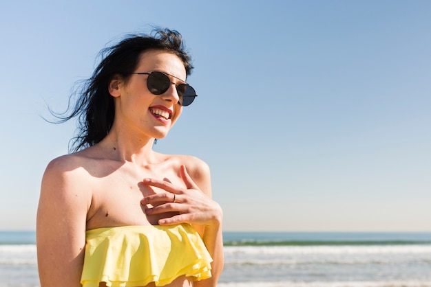 Porträt einer lächelnden jungen Frau in der Bikinioberseite, die gegen blauen Himmel am Strand steht