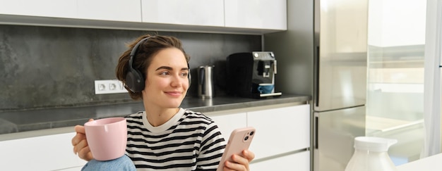 Kostenloses Foto porträt einer lächelnden jungen frau mit mobiltelefon, die in der küche sitzt und heißen tee trinkt