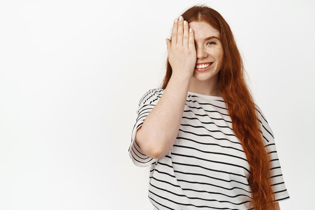 Porträt einer lächelnden, kurvigen Frau mit langen, natürlichen roten Haaren, Ingwerfrisur und Sommersprossen, bedeckt eine Seite des Gesichts mit einer Handfläche, schaut glücklich in die Kamera und steht vor weißem Hintergrund.