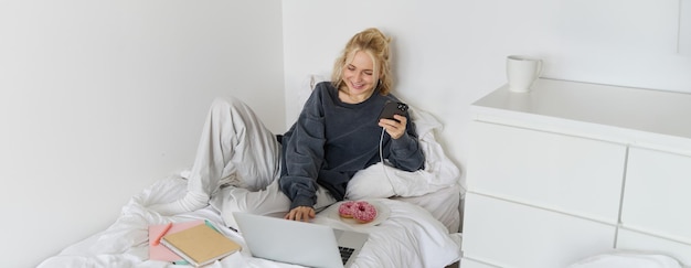 Kostenloses Foto porträt einer lächelnden, offenen frau, die mit einem smartphone und einem laptop im bett mit einem donut liegt