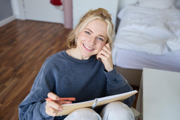 Porträt einer lächelnden schönen jungen ehrlichen Frau sitzt im Schlafzimmer mit Stift und Notizbuch schreiben in