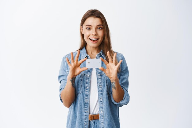 Porträt einer lächelnden und überraschten jungen Frau, die eine Plastikkreditkarte in der Hand hält, überreicht der Brust die Werbung für eine neue Bankfunktion, die weißen Hintergrund der Bank empfiehlt