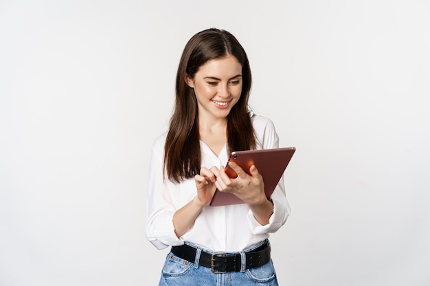 Porträt einer lächelnden Unternehmensfrau, die ein digitales Tablet betrachtet, arbeitet und über weißem Hintergrund steht.