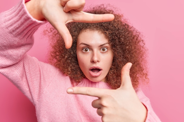 Kostenloses Foto porträt einer lockigen jungen frau sieht schockiert aus rahmen mit fingern sucht perfekte stelle oder perspektive misst winkel trägt lässigen pullover einzeln über rosafarbener wand. foto machen