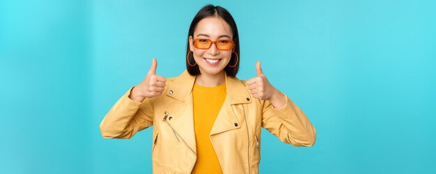 Porträt einer modernen jungen asiatischen Chinesin mit Sonnenbrille trägt trendige Oberbekleidung, die Daumen nach oben zeigt und erfreut lächelt und etwas blauen Hintergrund empfiehlt