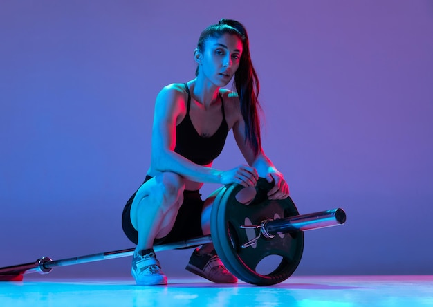 Kostenloses Foto porträt einer muskulösen frau, die mit einer langhantel trainiert, isoliert auf violettem hintergrund im neonlicht