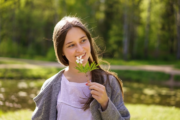 Porträt einer niedlichen glücklichen Frau auf dem Hintergrund eines Sees, eine Blume an ihrem Gesicht an einem sonnigen Frühlingstag haltend