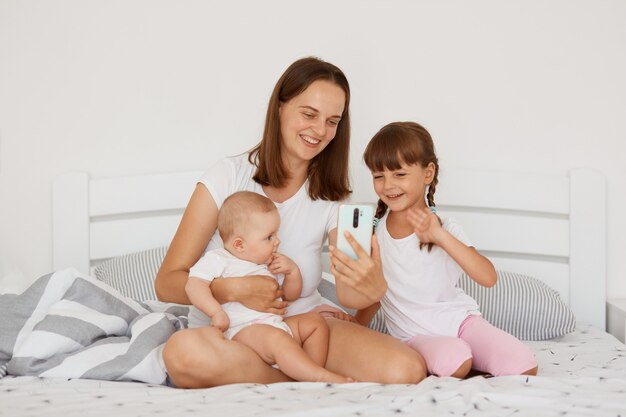 Porträt einer positiven Frau, die ein weißes T-Shirt im Casual-Stil trägt, das mit zwei Töchtern auf dem Bett sitzt, ein Smartphone in den Händen hält, einen Videoanruf hat oder einen Livestream sendet.
