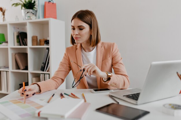 Porträt einer positiven Frau in einem stilvollen Geschäftsoutfit, das Dokumente auf ihrem Desktop mit Interesse betrachtet.