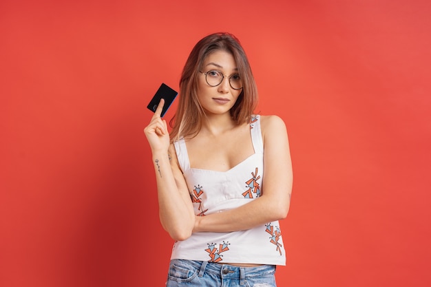 Porträt einer reizenden jungen Frau, die eine Brille trägt, die Kreditkarte auf roter Wand zeigt
