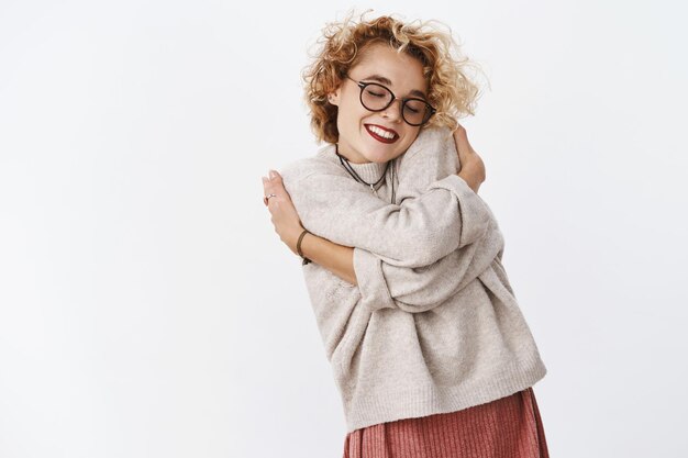 Porträt einer sanften und schönen schönen Frau in Pullover und Brille, die sich auf die Schulter stützt, umarmt sich mit den Händen über den Körper und lächelt sinnlich und fühlt sich wohl.