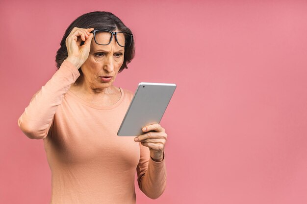 Porträt einer schockierten, erstaunten, schönen seniorin im alter, die einen tablet-computer verwendet, einzeln auf rosafarbenem hintergrund. kopieren sie platz für text.