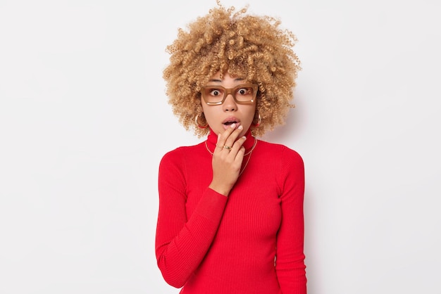 Kostenloses Foto porträt einer schockierten jungen frau mit lockigem haar keucht von wunderständen sprachlos hält den atem an kann nicht glauben, dass ihre augen eine brille tragen und der rote rollkragenpullover auf etwas atemberaubendes reagiert.