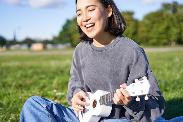 Porträt einer schönen asiatischen Frau, die im Park singt und Ukulele-Gitarre spielt und allein auf Gras sitzt