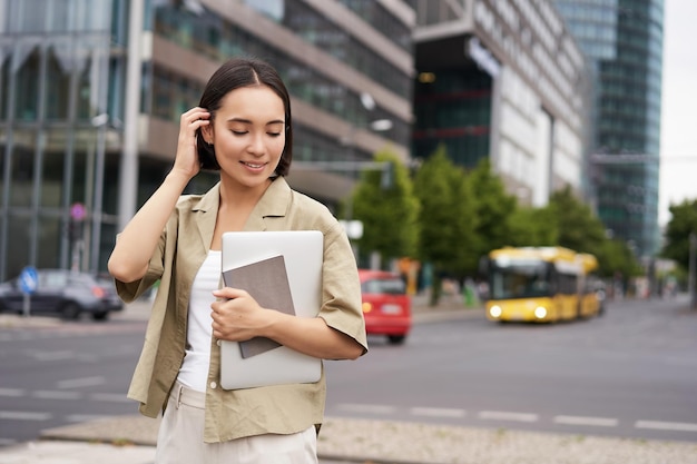 Porträt einer schönen asiatischen Frau, die mit Laptop und Notizbuch auf der Straße steht und zur Universität geht oder