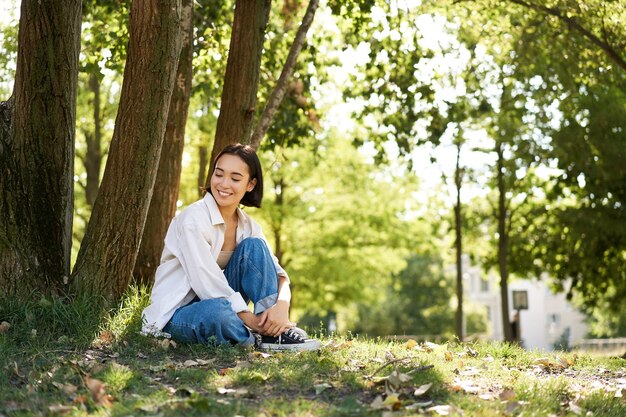 Porträt einer schönen asiatischen Frau, die sich in der Nähe eines Baumes ausruht und sich im Park entspannt, lächelnd und glücklich aussieht