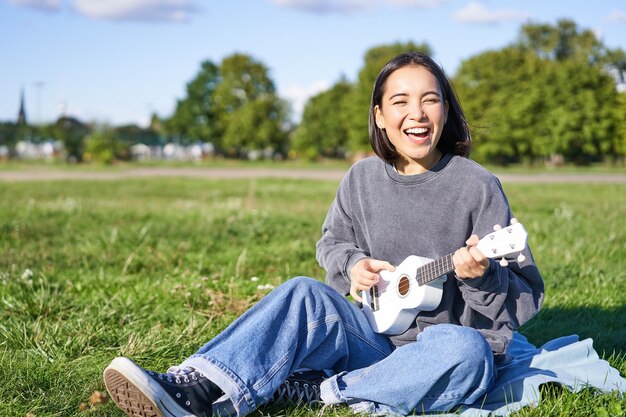 Porträt einer schönen asiatischen Frau, die singt und Ukulele-Gitarre im Park spielt, allein sitzend auf Gras auf s