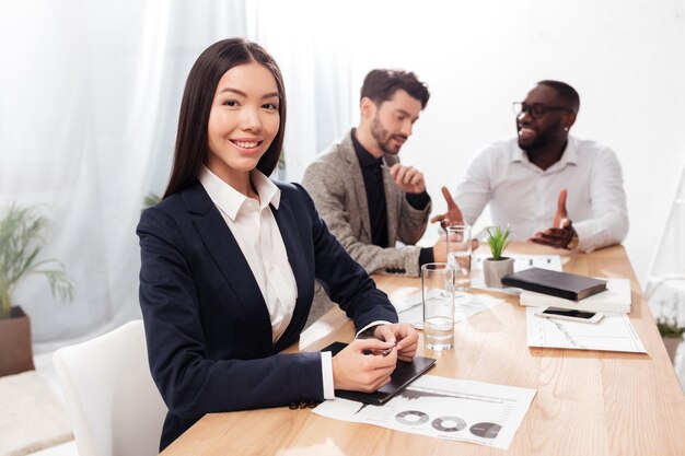 Porträt einer schönen asiatischen Geschäftsfrau, die im Büro sitzt und glücklich mit multinationalen Geschäftspartnern im Hintergrund in die Kamera blickt