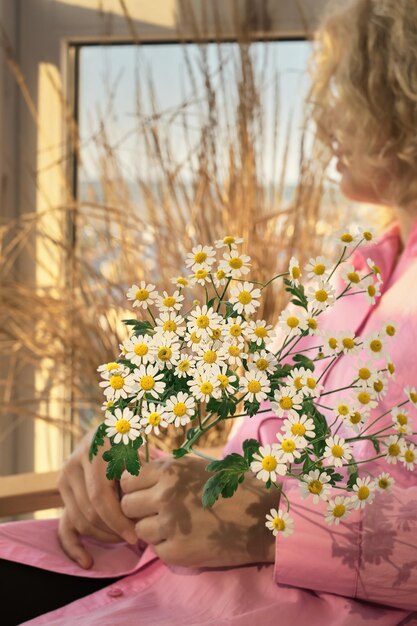 Porträt einer schönen blonden erwachsenen Frau mit einem Blumenstrauß aus Gänseblümchen sitzt auf einem Stuhl und sieht weg Nahaufnahme selektiver weicher Fokus auf Blumen vertikalen Rahmen