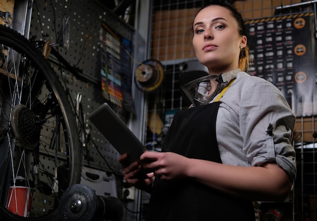 Porträt einer schönen brünetten Frau, die Arbeitskleidung, Schürze und Schutzbrille trägt, hält einen Tablet-Computer in einer Werkstatt