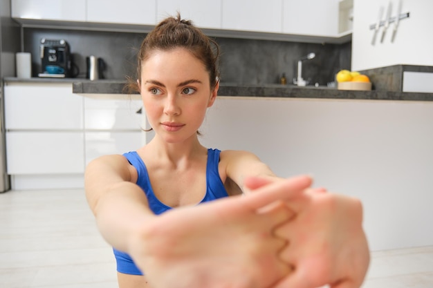Kostenloses Foto porträt einer schönen frau beim fitnesstraining, sitzt auf dem boden in der nähe der küche und streckt ihre hände beim training von zu hause aus kopierraum