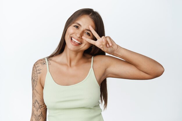 Porträt einer schönen Frau mit Tätowierungen, die ein Friedenszeichen gegen das Auge zeigen und glücklich lächeln, über weißem Hintergrund stehen.
