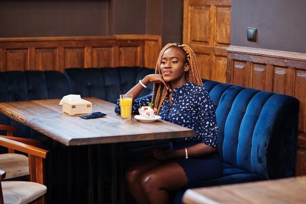 Porträt einer schönen jungen afrikanischen Geschäftsfrau mit Dreadlocks auf blauer Bluse und Rock, die im Café mit Eis und Ananassaft sitzt