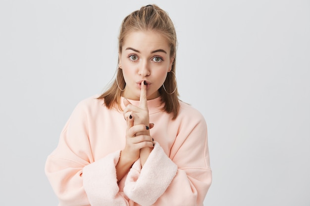 Porträt einer schönen jungen blonden Frau, die ein stilvolles rosa Sweatshirt trägt und darum bittet, still zu bleiben oder ihre Geheimnisse nicht zu verraten, einen ernsten Blick zu haben und den Finger auf ihren Lippen zu halten. Körpersprache