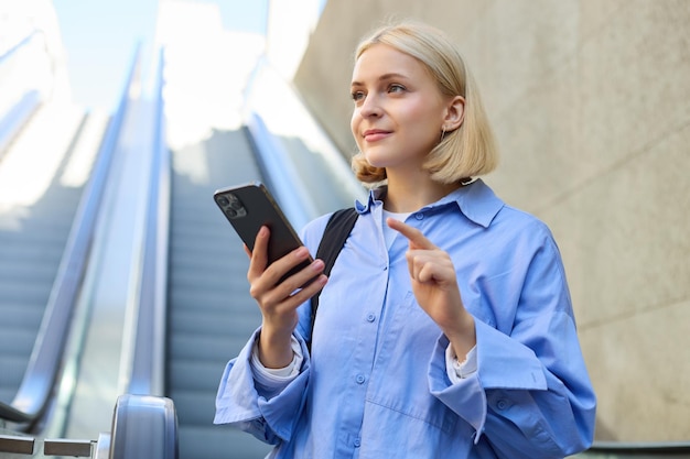 Kostenloses Foto porträt einer schönen jungen blonden frau in blauem hemd, die in der nähe der rolltreppe steht und ein mobiltelefon in der hand hält