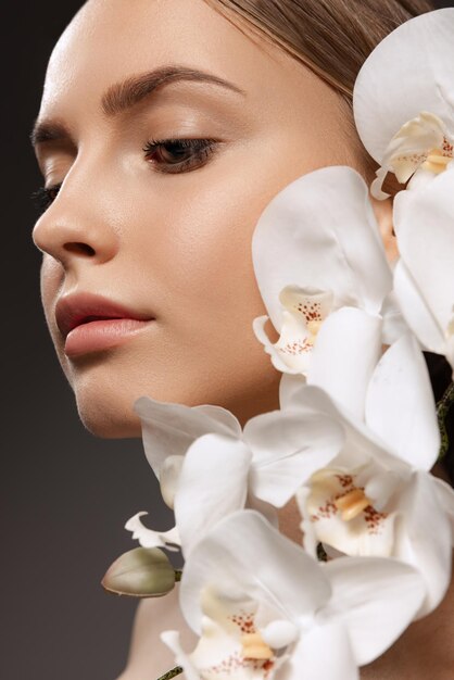 Porträt einer schönen jungen Frau mit nacktem Make-up, die mit Orchideenblüten einzeln auf dunklem Hintergrund posiert