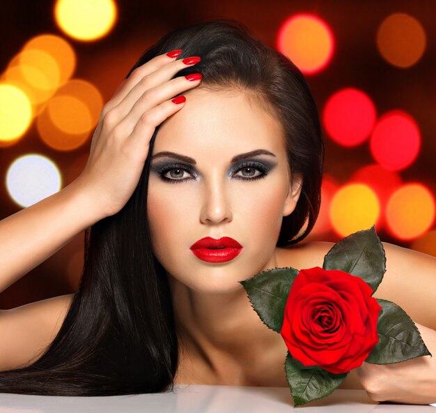 Porträt einer schönen jungen Frau mit roten Lippen, Nägeln und Rosenblume in der Hand. Modemodell mit dem Make-up des blauen Auges, das im Studio über Nachtlichtkugeln aufwirft. Weiches Bokeh-Hintergrundkonzept.