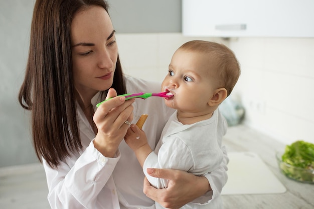 Porträt einer schönen jungen Mutter, die ihr neugeborenes Kind mit Brei füttert Sohn, der vom Löffel isst und mit lustigem Ausdruck zur Seite schaut