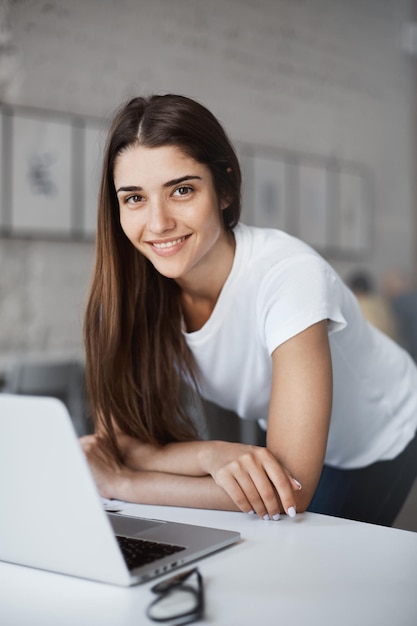 Porträt einer schönen kaukasischen Frau, die einen Laptop verwendet, der im Coworking Center im offenen Raum steht und nach einem neuen Job sucht, der lächelnd in die Kamera schaut