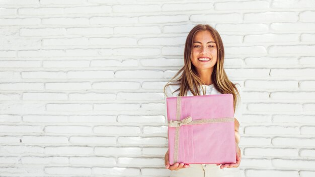 Porträt einer schönen lächelnden Frau, die rosa Geschenkbox gegen Backsteinmauer hält