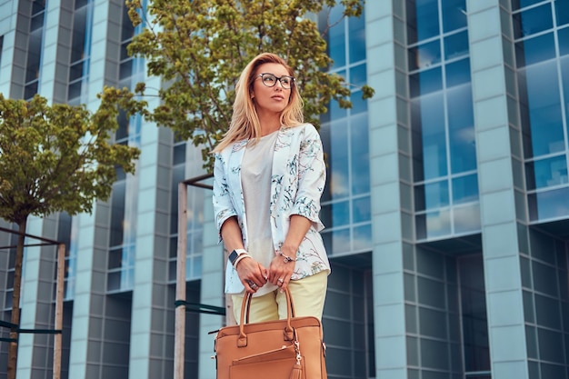 Porträt einer schönen modischen Frau in stilvoller Kleidung und Brille mit Handtasche, die vor einem Wolkenkratzer steht.