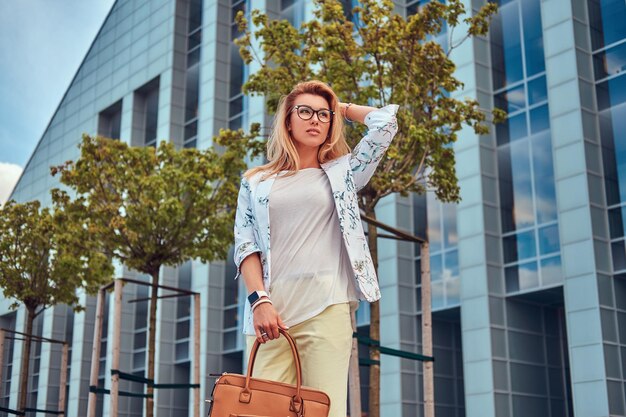 Porträt einer schönen modischen Frau in stilvoller Kleidung und Brille mit Handtasche, die vor einem Wolkenkratzer steht.