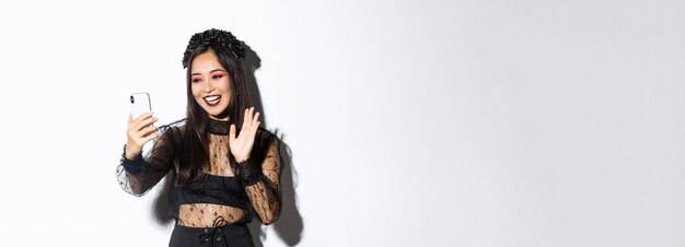 Porträt einer schönen und stilvollen asiatischen Frau im gotischen Spitzenkleid, die Smartpho mit der Hand winkt