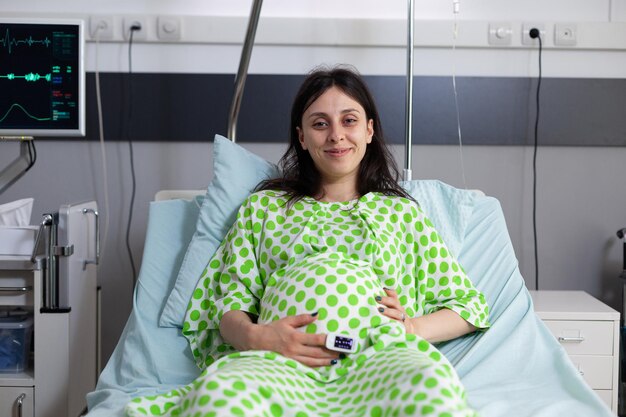 Porträt einer schwangeren Frau, die im Krankenbett sitzt und in die Kamera schaut. Junge Person, die lächelt, auf die Geburt des Kindes und die Elternschaft in der medizinischen Klinik wartet. Erwachsener mit Babybauch