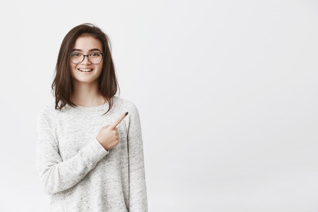 Porträt einer sehr glücklichen jungen Frau in der Brille, die auf den oberen rechten Conner zeigt, während sie breit lächelt