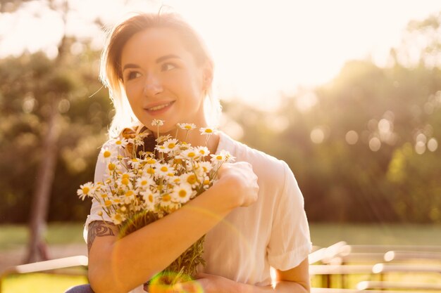 Porträt einer sonnigen Frau, die einen Haufen Kamille mit den Sonnenstrahlen auf ihrem Gesicht umarmt und das Leben genießt