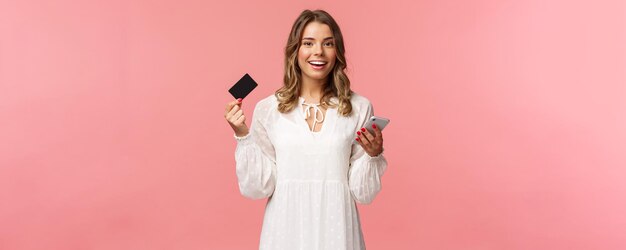 Porträt einer sorglosen jungen hübschen blonden Frau, die mit dem Handy für den Online-Kauf mit Kreditkarte bezahlt und glücklich lächelt