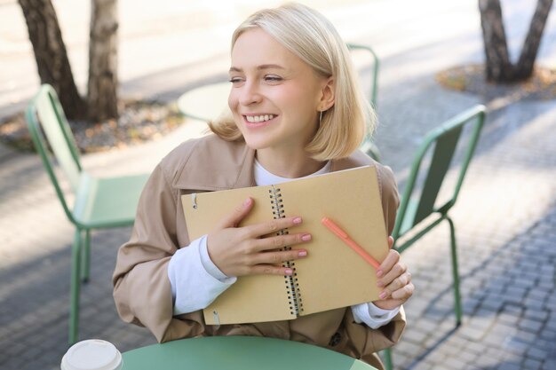 Kostenloses Foto porträt einer sorglosen lächelnden jungen frau mit notizbuch und stift, die in einem café im freien in der nähe eines tisches sitzt