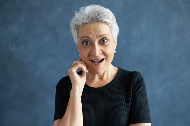 Porträt einer stilvollen attraktiven Frau im Ruhestand mit grauem Haar, das den Mund öffnet und aufgeregt ausruft, Erstaunen ausdrückt, von unerwarteten Neuigkeiten überrascht ist und die Hand an ihrem Gesicht hält