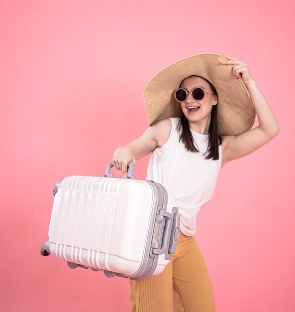 Porträt einer stilvollen jungen Frau in Sommerkleidung und einem Weidenhut mit einem Koffer auf einem isolierten Rosa