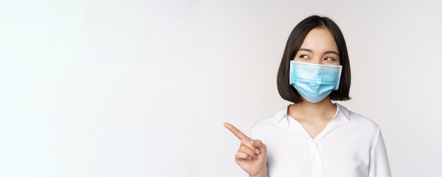 Porträt einer süßen asiatischen Frau mit medizinischem Gesichtsmasken-Coronavirus-Schutz, die mit dem Finger nach links und lo zeigt